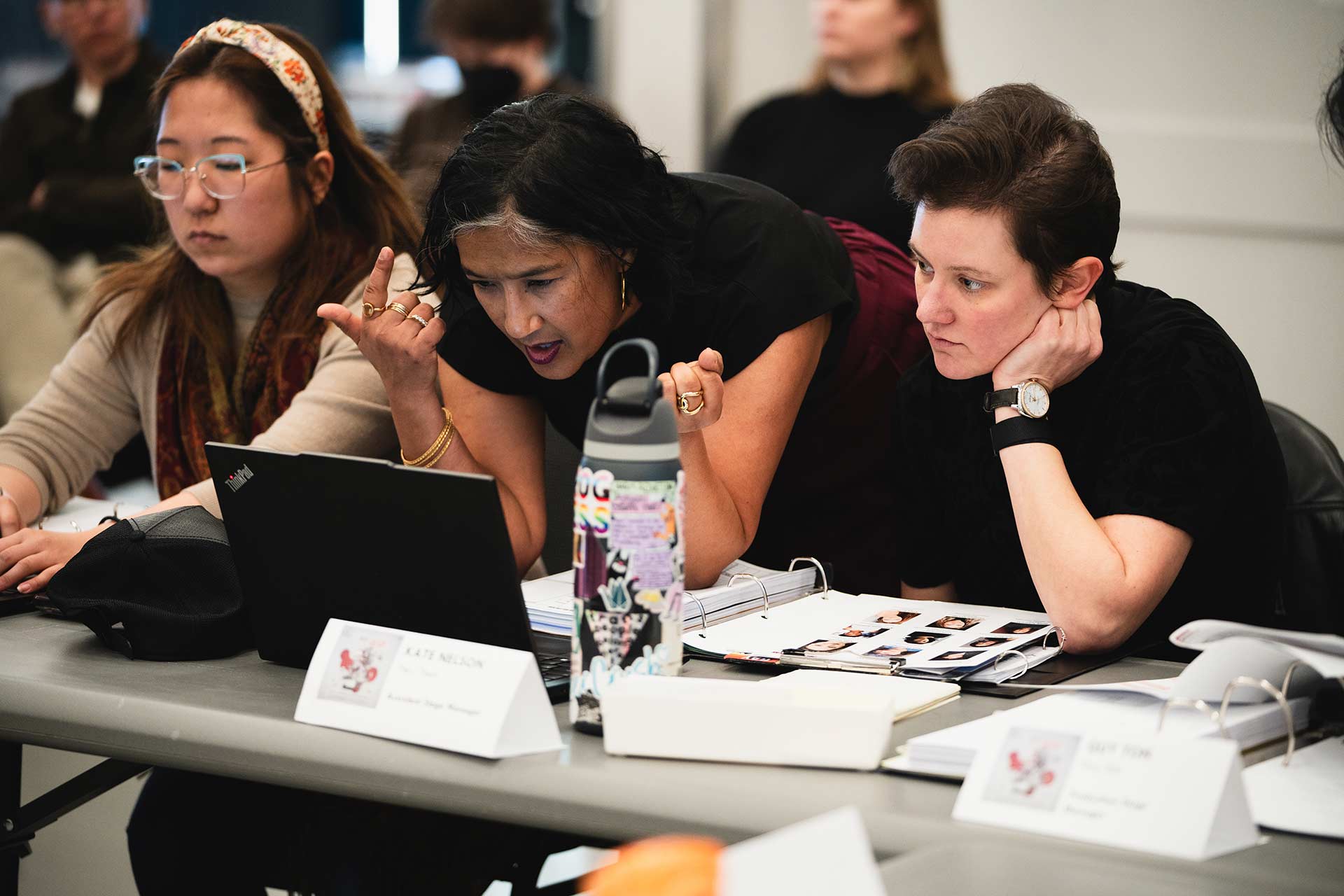 Three individuals sitting at a table covered with papers and a laptop. The person in the middle leans over, appearing to explain something on the screen to the others. Water bottles, folders, and name tags are on the table.