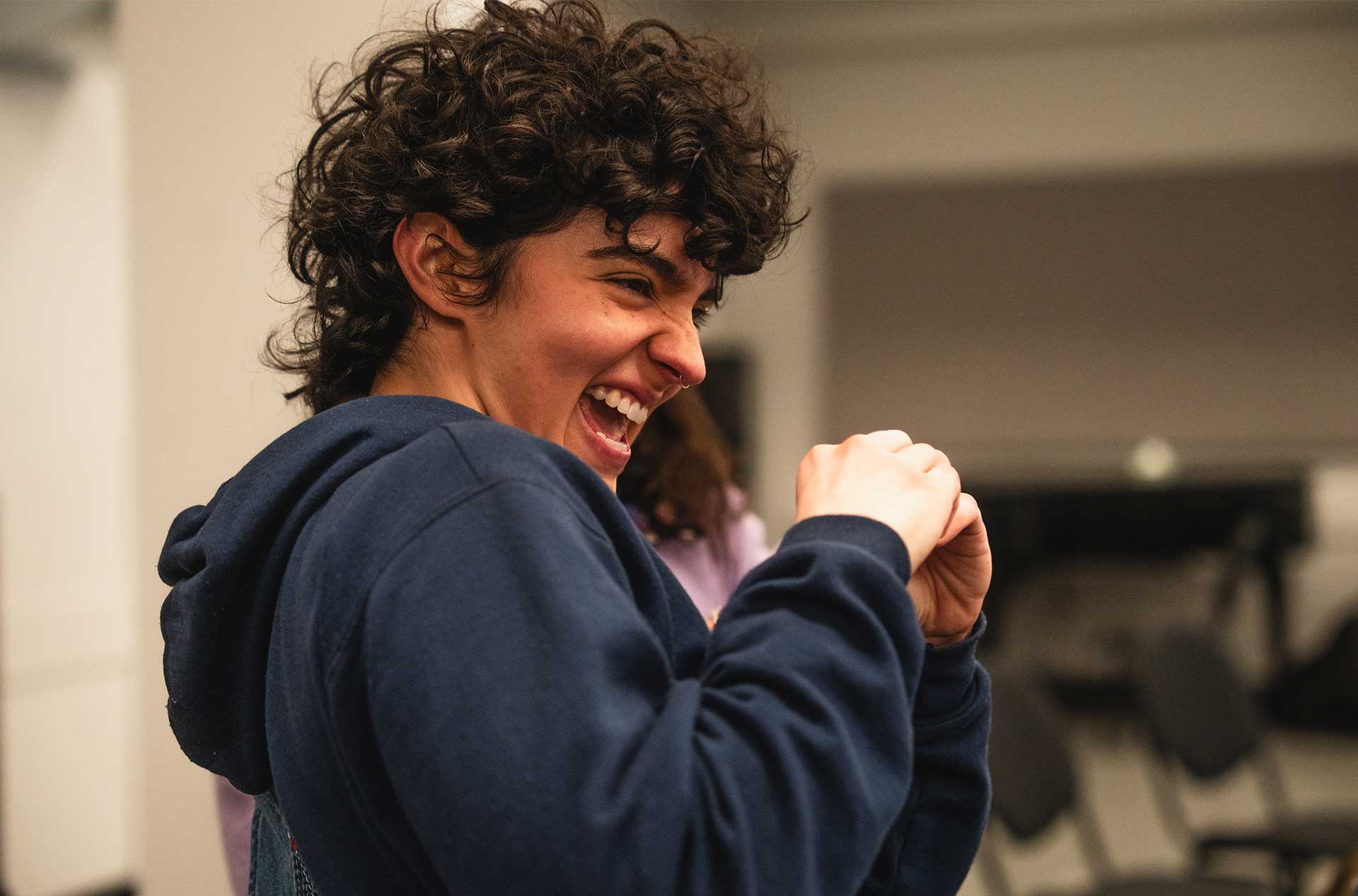 A person with short curly hair laughs joyfully while mid-gesture, standing in a rehearsal space.