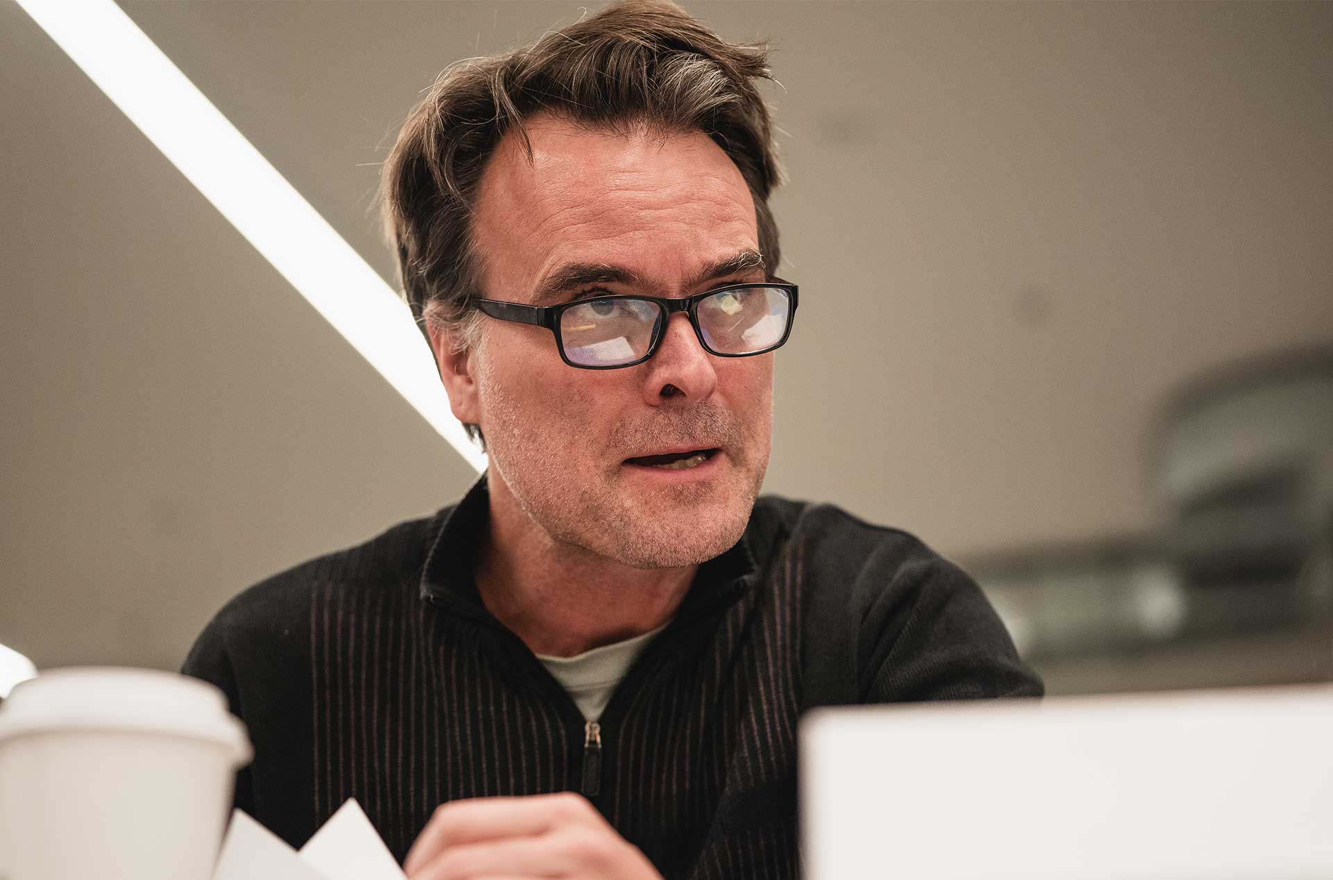 A person with short, tousled hair and glasses looks focused while speaking during rehearsal, with a coffee cup and script in front.