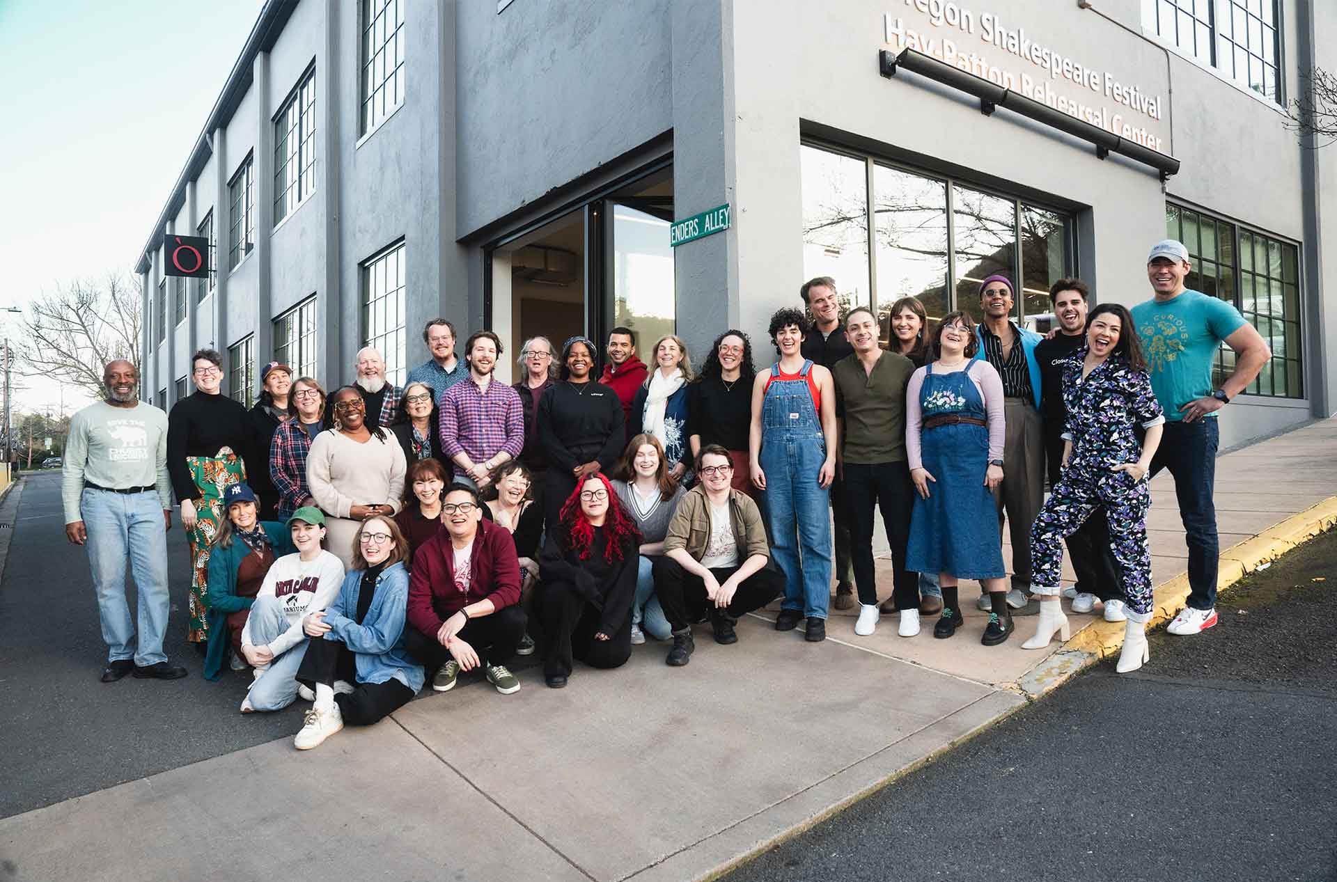 A large group stands smiling outside the Oregon Shakespeare Festival Hay-Patton Rehearsal Center, posing for a group photo.