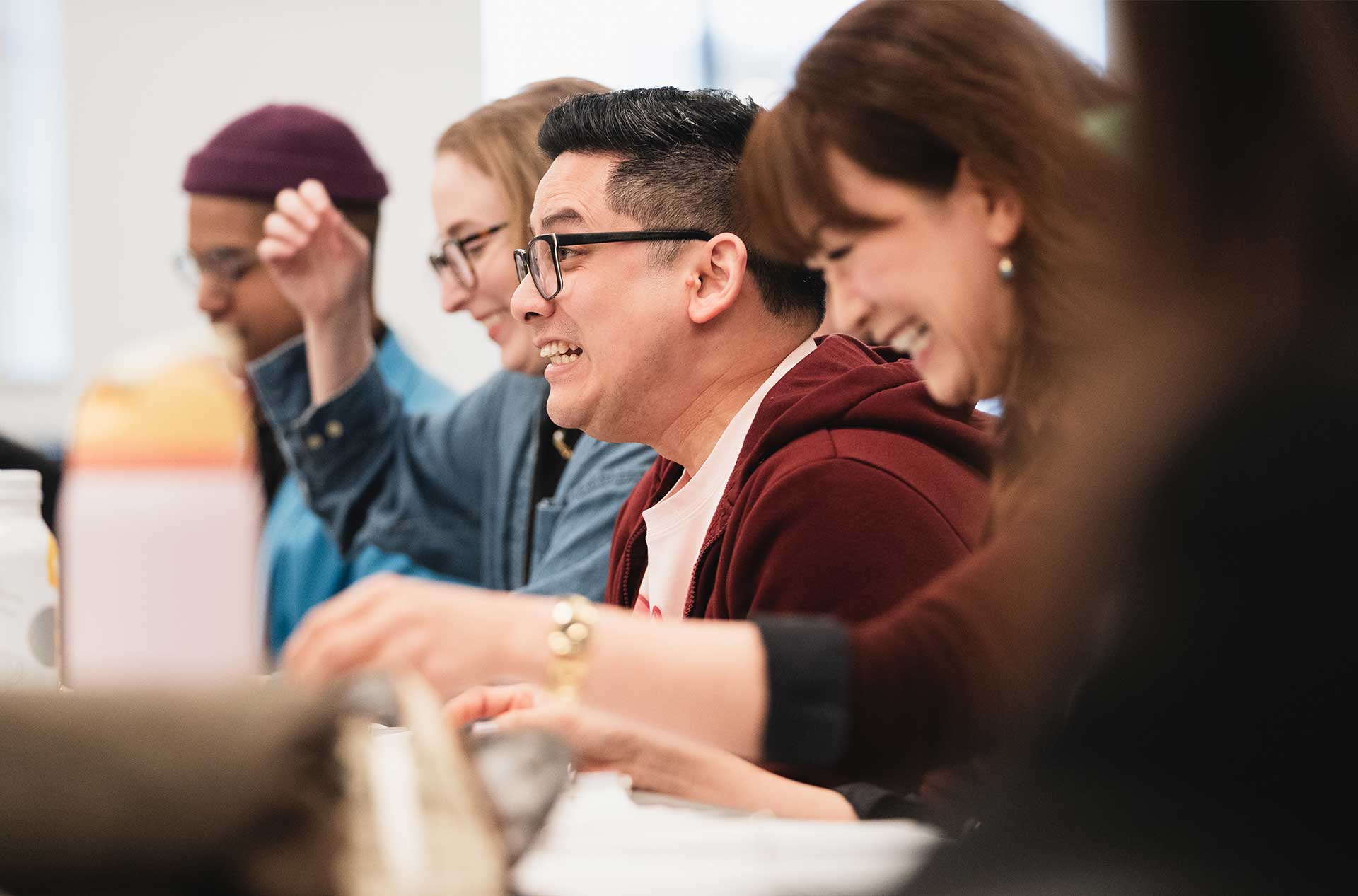 A group seated side by side at a rehearsal table reacts with laughter and wide smiles, immersed in the moment.