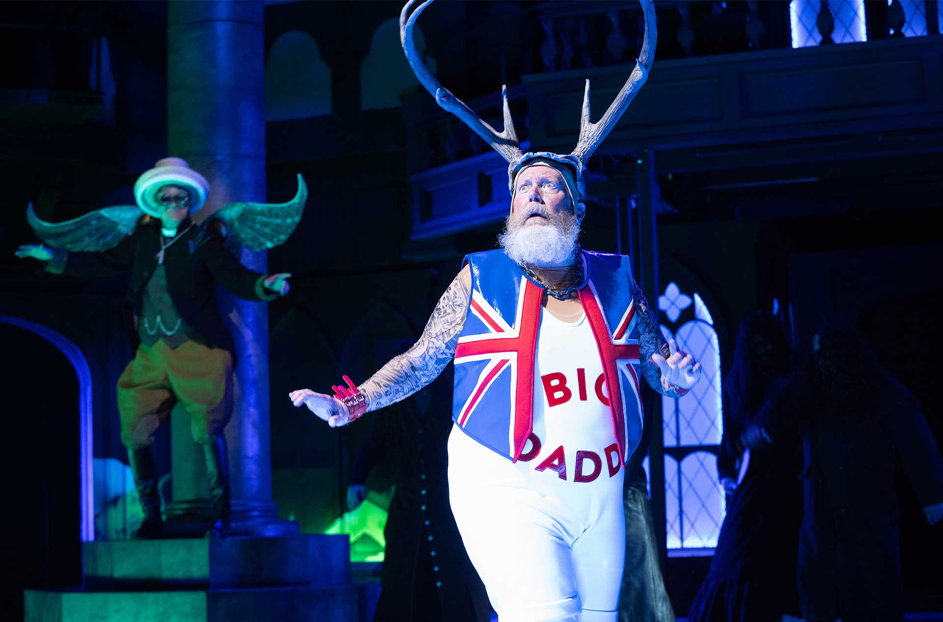 A white-bearded actor wearing a Union Jack vest over a bodysuit labeled “BIG DADDY” and large antlers on their head looks wide-eyed, while another character with wings stands behind on a platform.