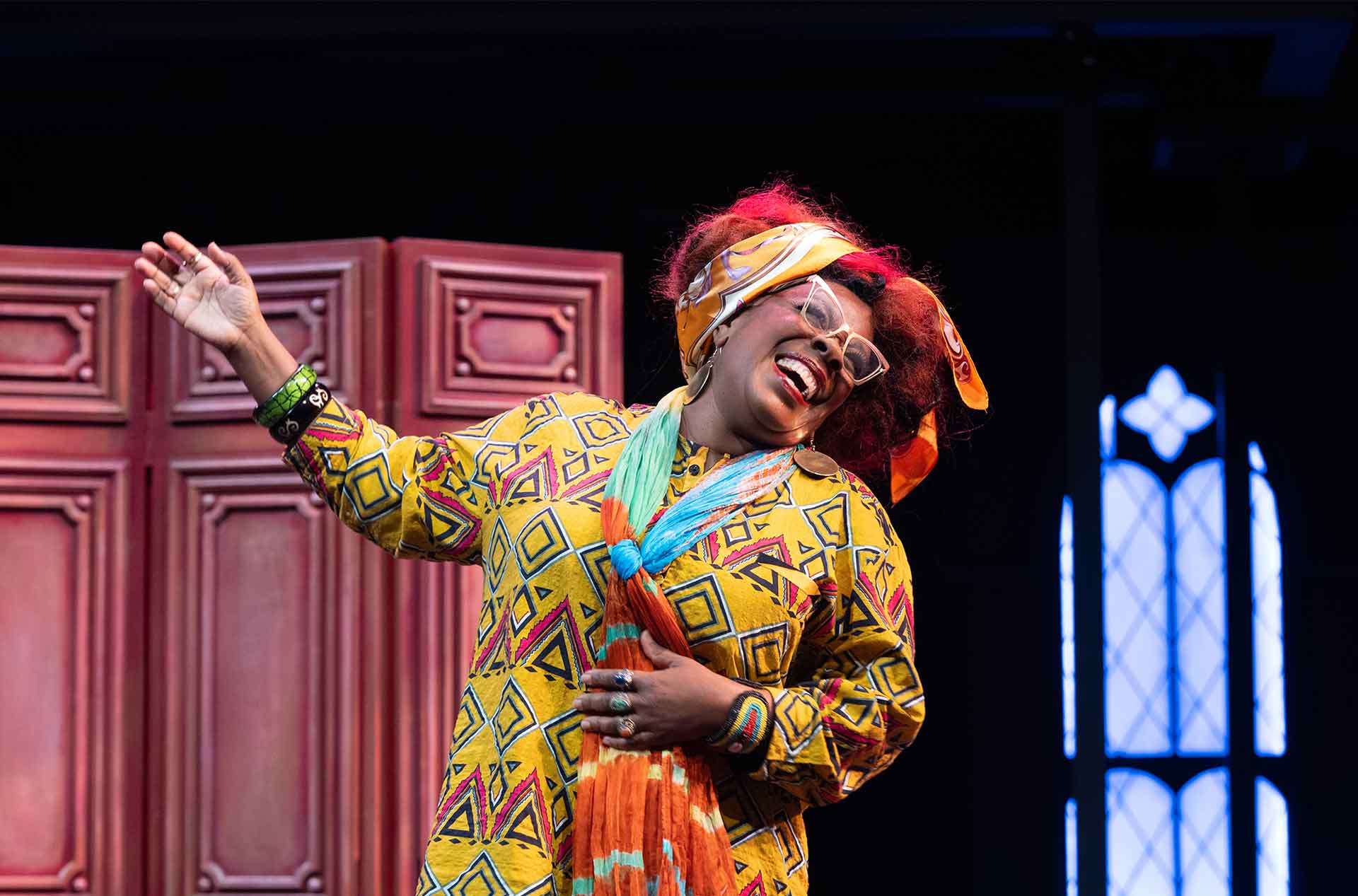 A joyful performer in a bright yellow patterned dress with geometric shapes and colorful scarf sings with arms open wide, standing in front of a red paneled screen and blue-lit stained glass window.