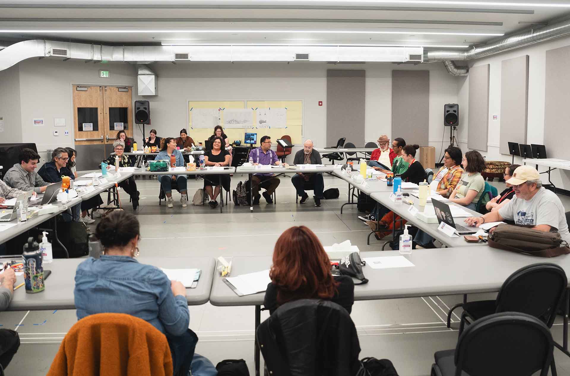 Wide view of a rehearsal room with participants seated around a large U-shaped table, engaged with scripts, laptops, and notes.