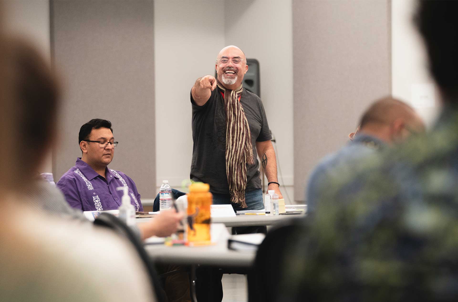 A person standing and smiling while pointing toward the camera during a group meeting, with others seated and focused around a table in a reheasal room.