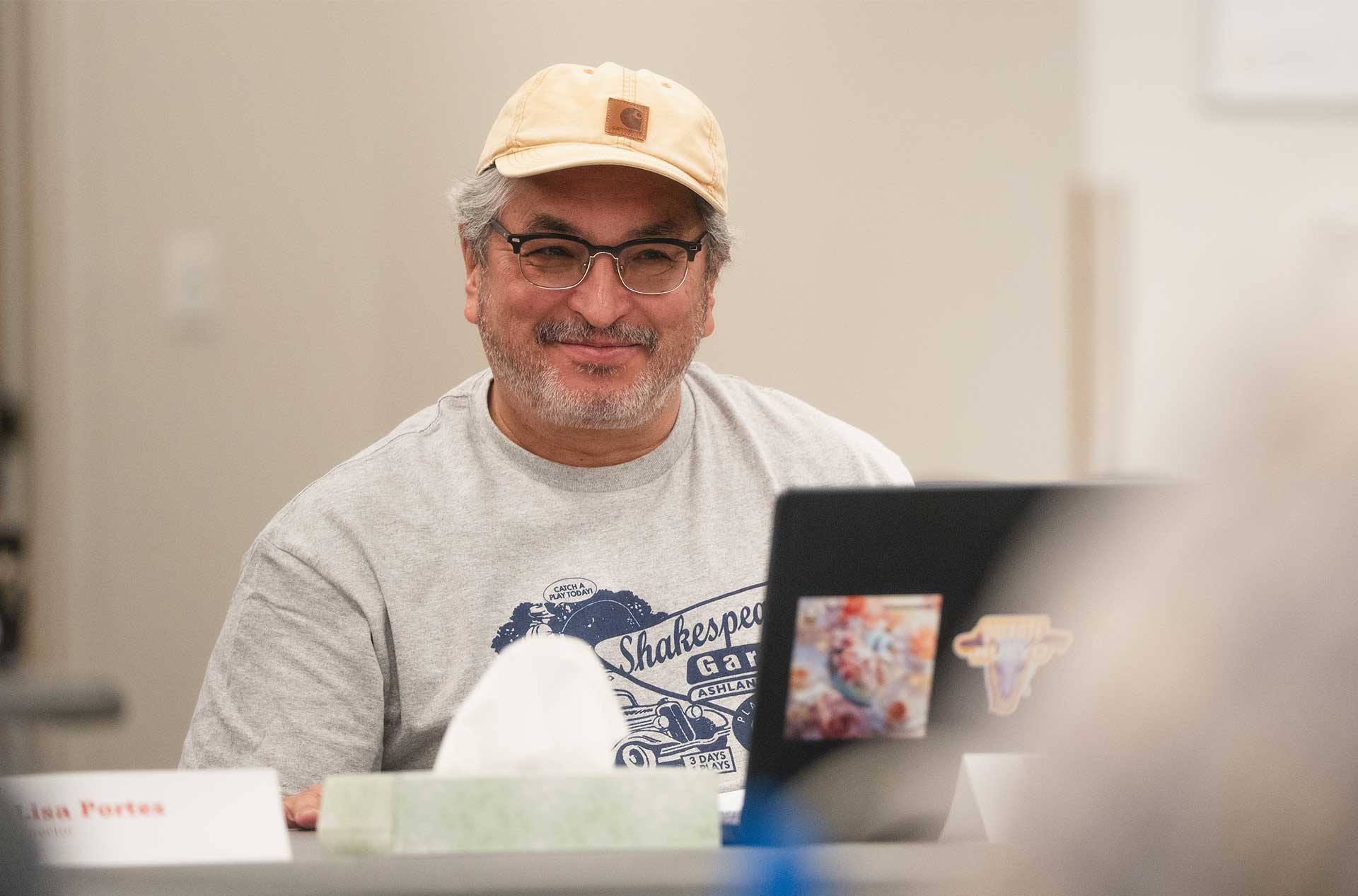 Person smiling while seated at a rehearsal table behind a laptop decorated with stickers, wearing glasses and a light-colored cap.