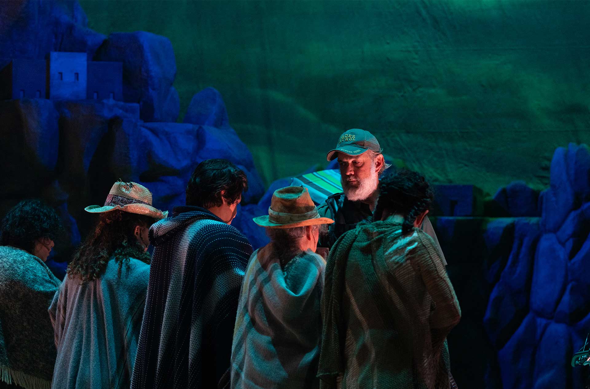 Group of actors in ponchos and hats surround a central figure in a “Border Patrol” cap, all set against a dark green and blue mountainous backdrop.