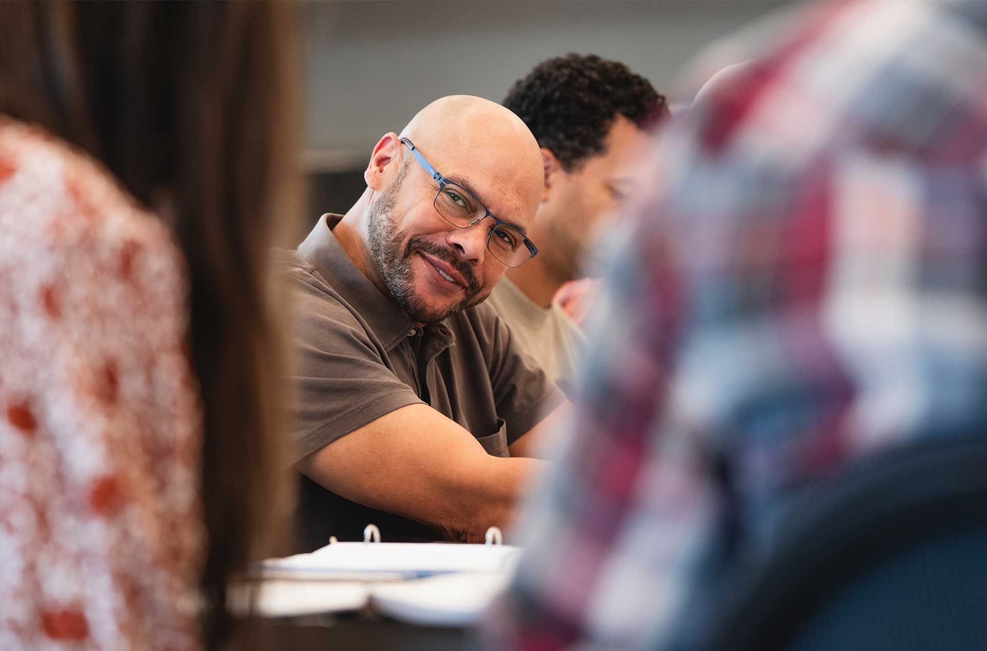 A person wearing glasses looks up and smiles during a conversation, seated at a table with open binders and surrounded by others out of focus.