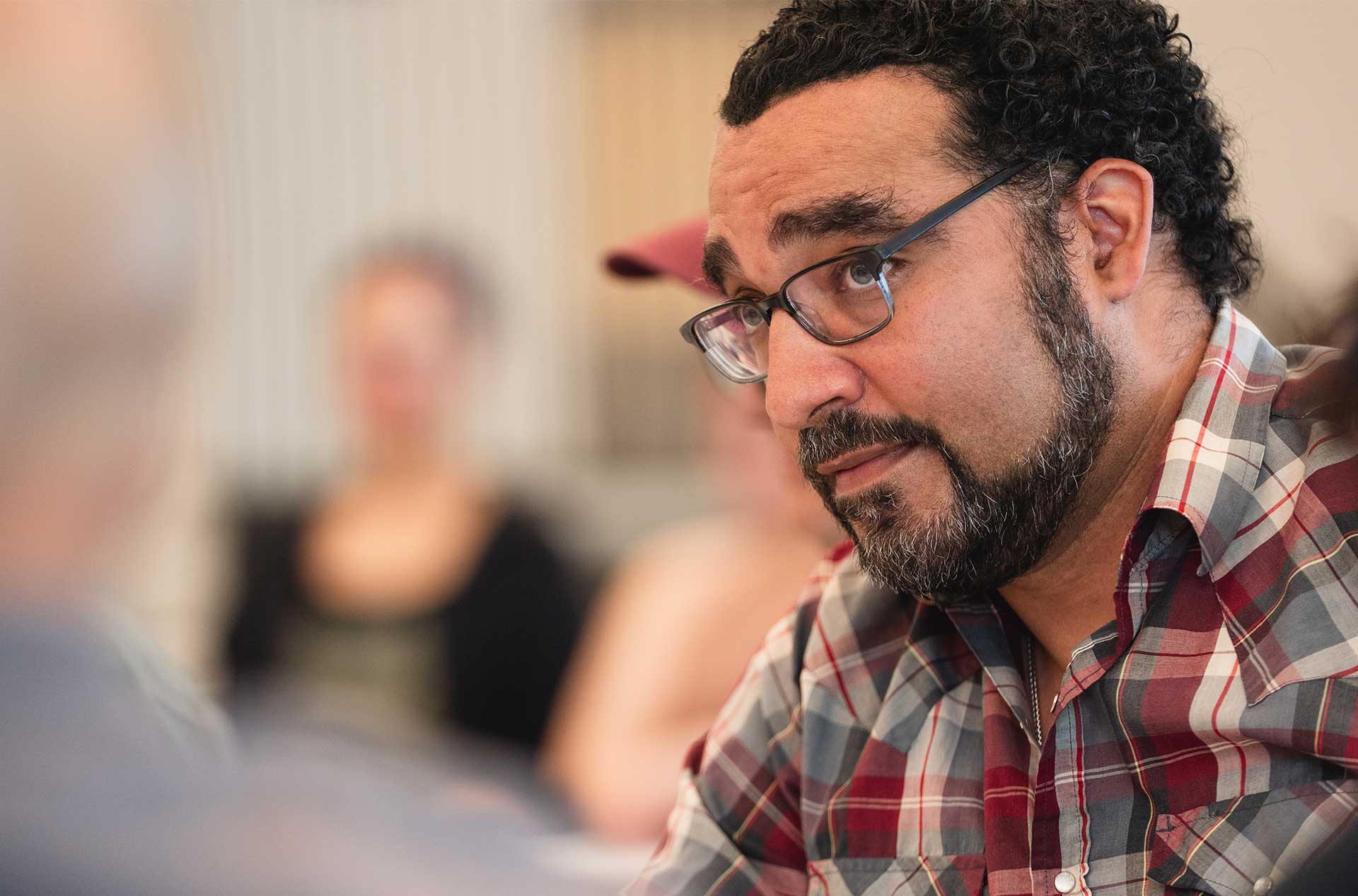 A close-up of a person with curly hair, glasses, and a plaid shirt listening intently, with blurred figures and a softly lit room in the background.