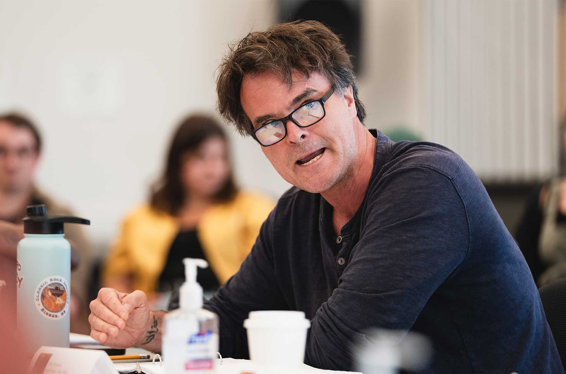 A person wearing glasses gestures while speaking at a table, with a water bottle, hand sanitizer, and coffee cup in the foreground.