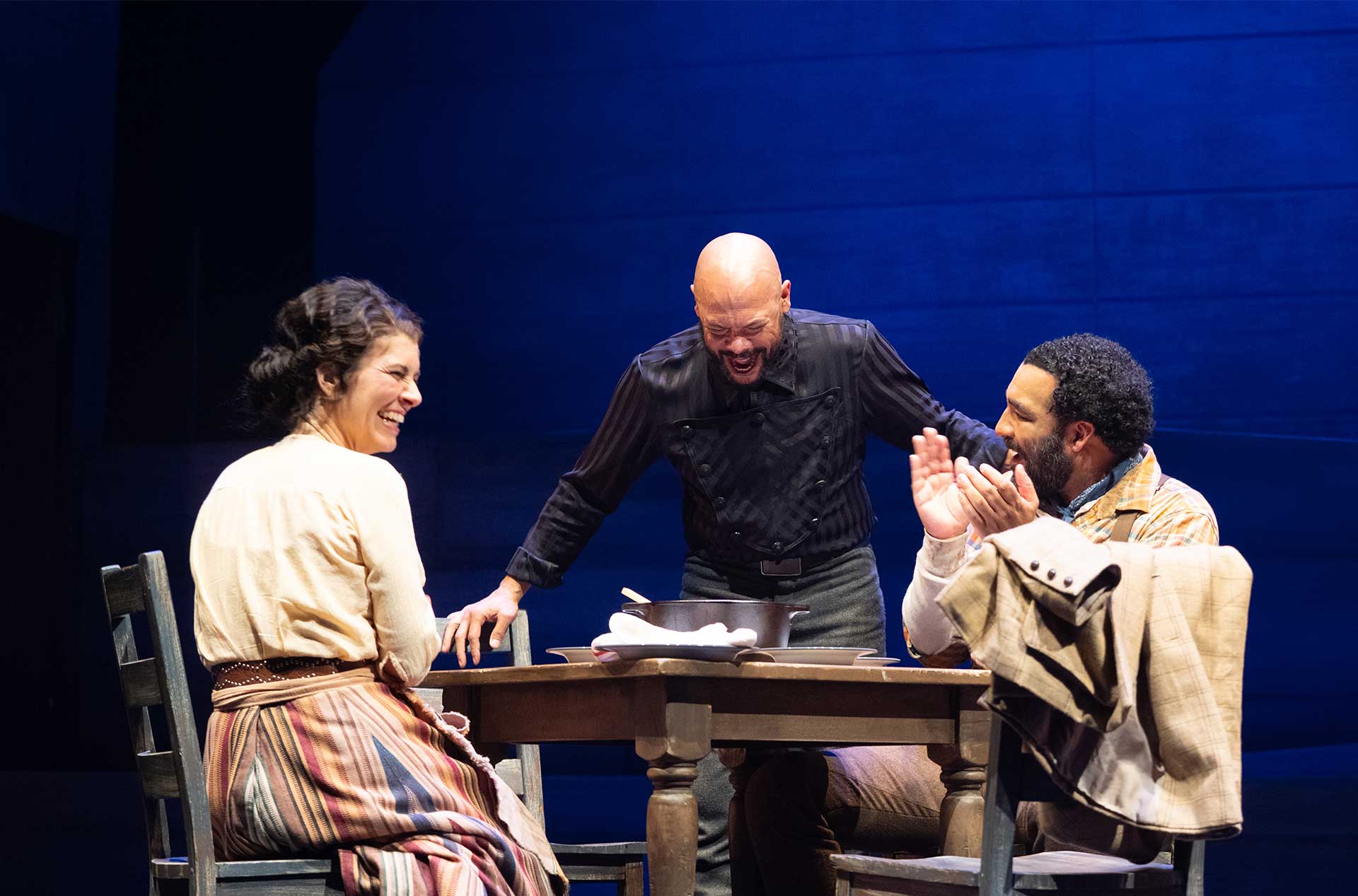 Three performers share a moment of laughter around a wooden table during a stage scene.