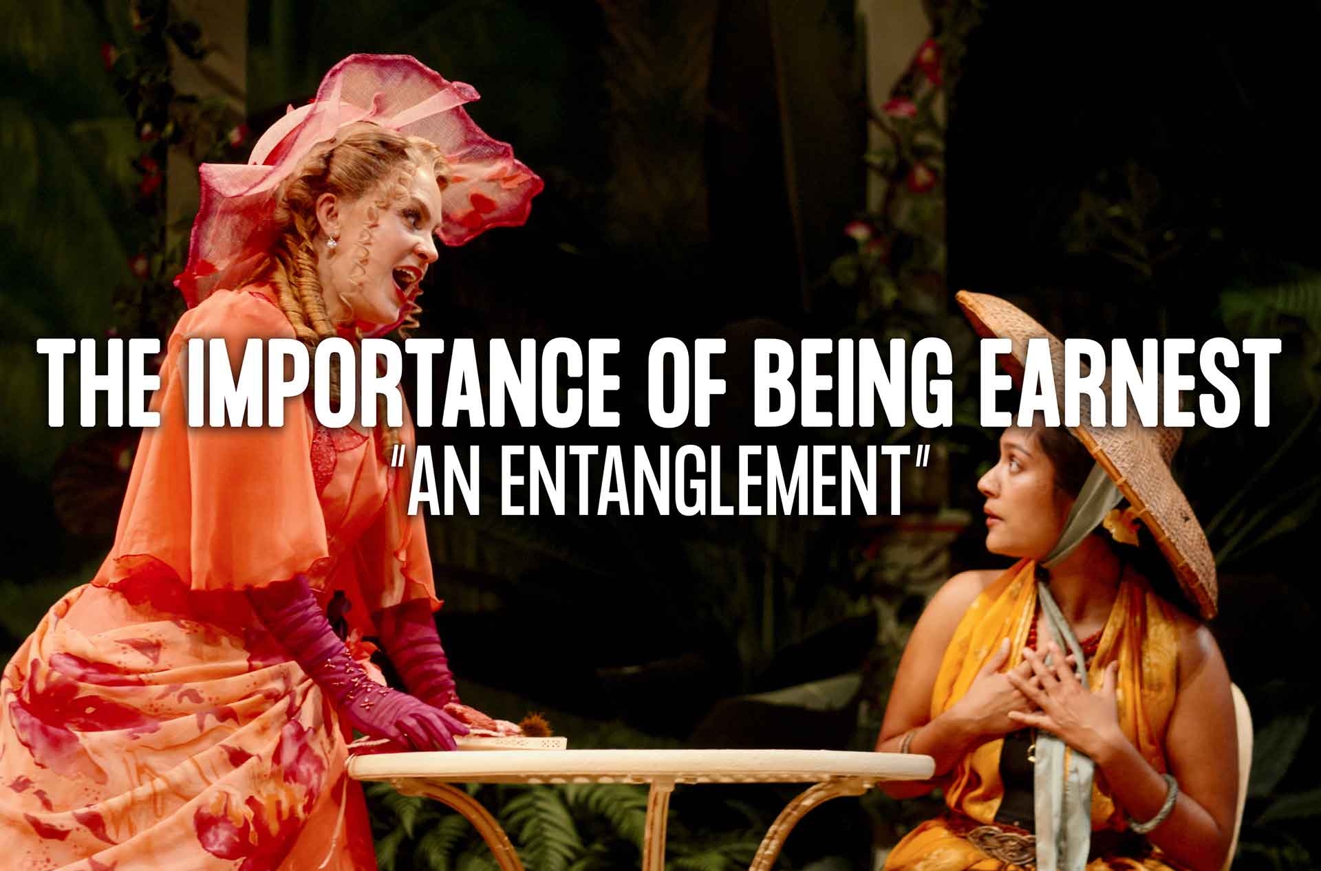 Two actors in colorful Victorian-style costumes perform a dramatic scene at a small garden table, surrounded by lush greenery.