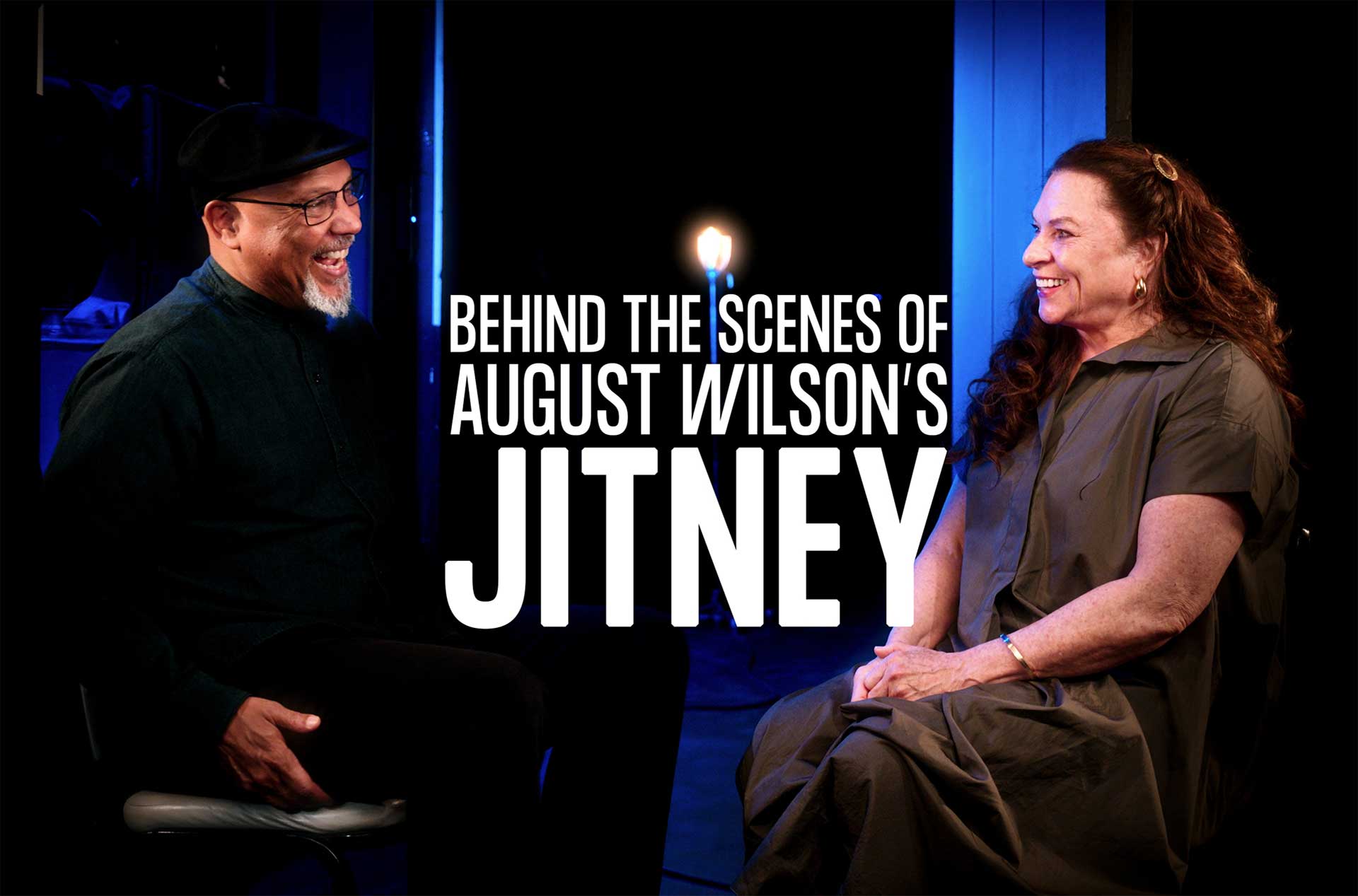 Two people are seated facing each other in a dimly lit space with a blue-toned background. They are engaged in conversation, smiling warmly. A single glowing lightbulb stands in the background, adding to the intimate atmosphere.