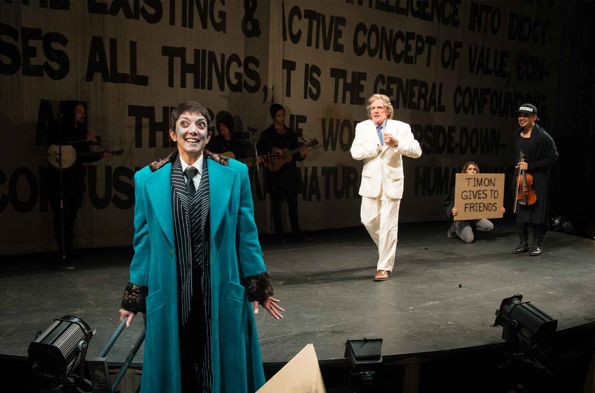 Person holding a sign with a person walking away from them and another person standing at the front of the stage looking ahead.