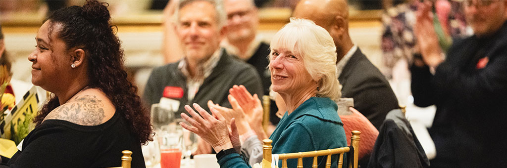 A group of people seated at round tables applauds during an indoor event, with one person smiling and looking toward the stage.