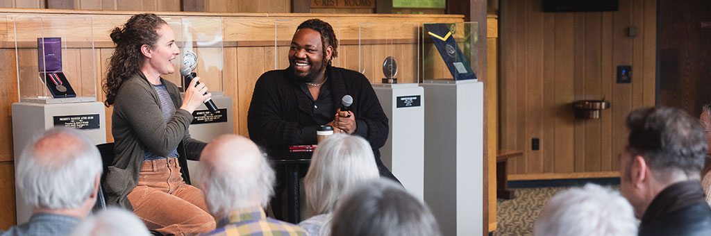 Two speakers holding microphones sit at the front of a room, smiling and engaging with an audience during a discussion.