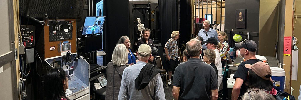 A group of visitors stands behind the scenes in a theater, surrounded by technical equipment and backstage structures.
