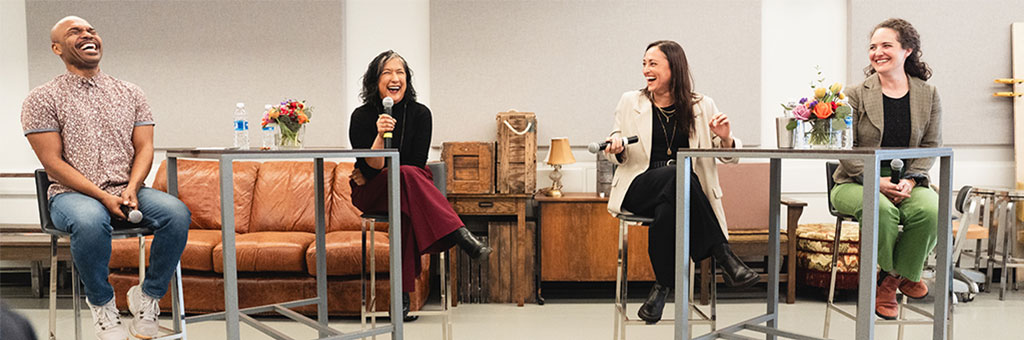 Four speakers sit at high tables in a casual panel setting, laughing and holding microphones in front of an audience.