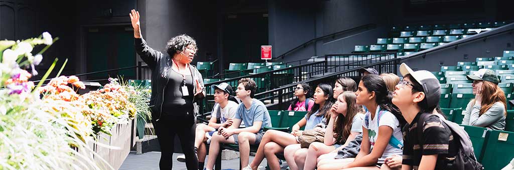 A group of people, mostly young students, sits attentively in an outdoor amphitheater while a guide speaks to them. The guide gestures with one hand, and the audience listens intently.