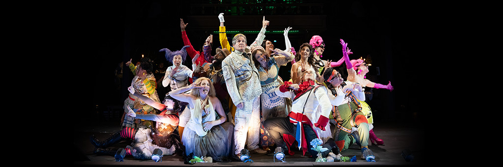 Performers in vibrant, eclectic costumes strike exaggerated poses on stage during a theatrical production, with dramatic lighting highlighting the ensemble.