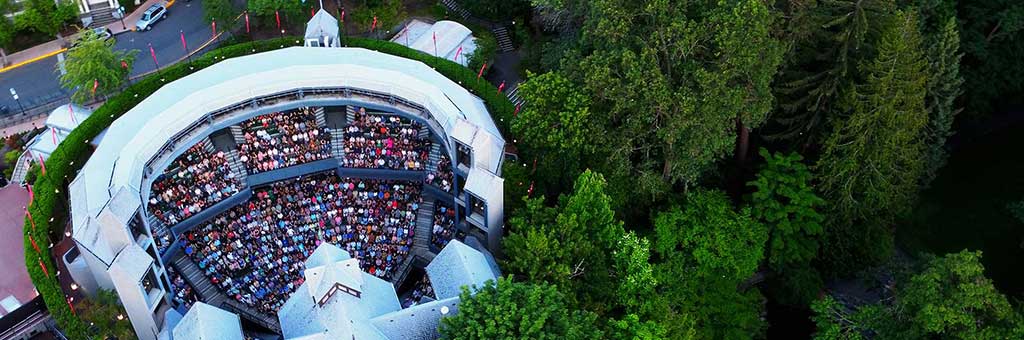 Aerial view of the Allen Elizabethan Theatre, showcasing its open-air seating filled with a large audience. The iconic semi-circular structure is surrounded by lush green trees, creating a picturesque setting.