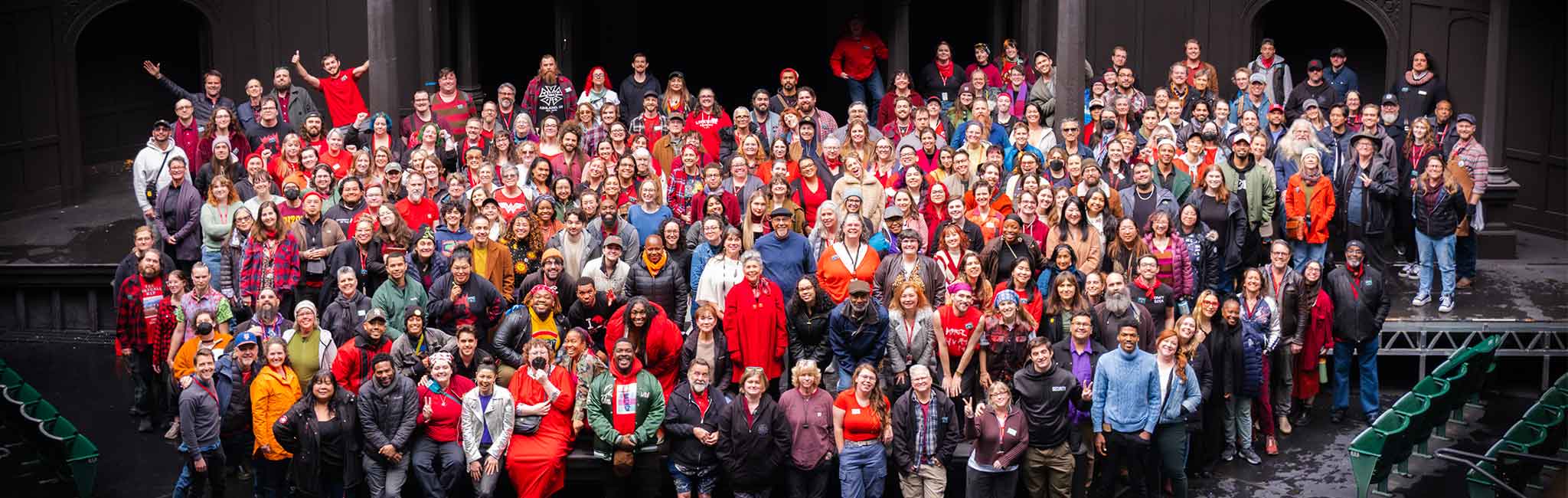 Close-up view of a large, diverse group of people smiling and posing together on a dark theater stage, framed by the Tudor-style backdrop.