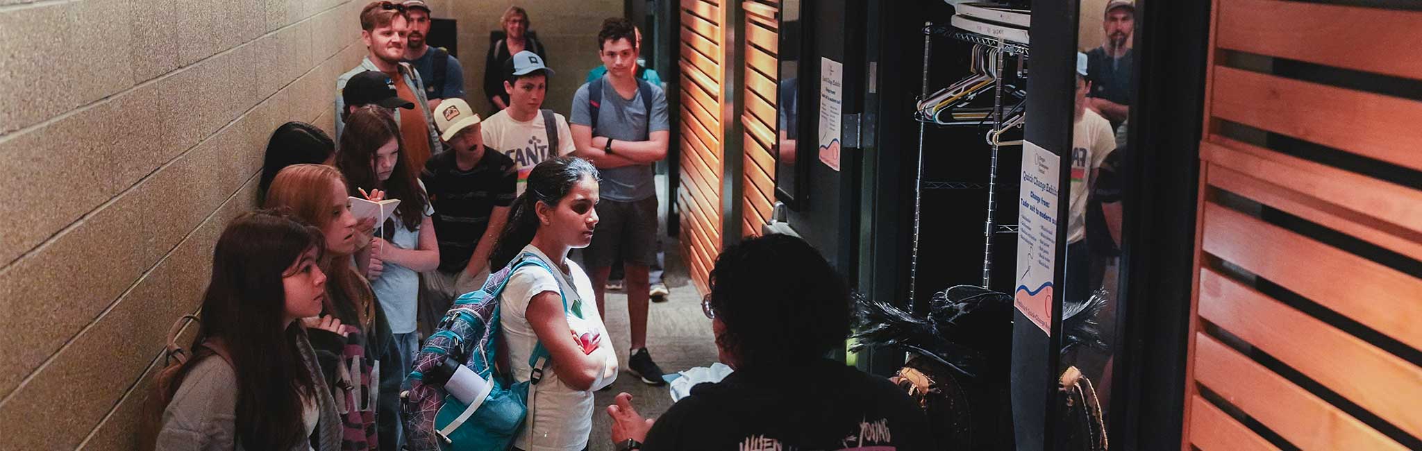 A group of people standing in a narrow hallway, attentively listening to someone at the front. The group appears engaged, some with backpacks and notepads, while looking at an open closet or storage area with hangers.