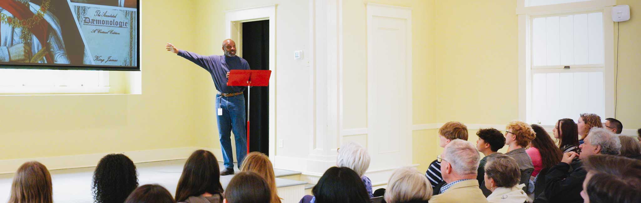 A speaker in a blue sweater gestures toward a projected slide featuring a royal figure and the book The Annotated Dæmonologie by King James. An attentive audience listens in a bright room with cream-colored walls and large windows.