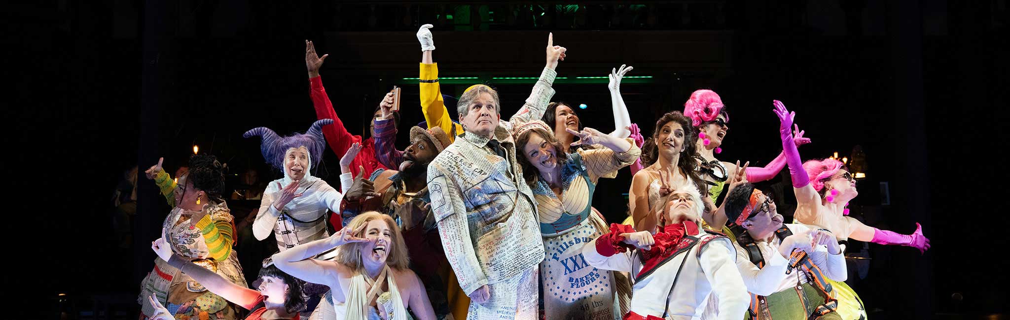 Performers in vibrant, eclectic costumes strike exaggerated poses on stage during a theatrical production, with dramatic lighting highlighting the ensemble.