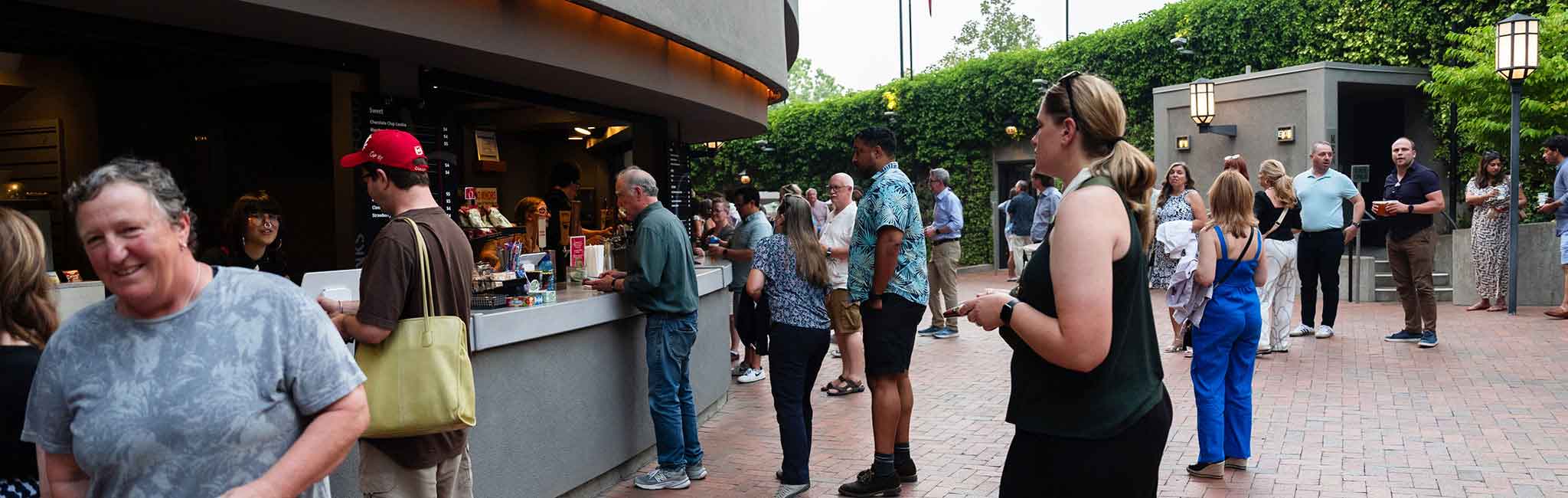 People gather in an outdoor courtyard, some ordering from a concessions stand while others socialize in small groups. The space is surrounded by greenery.