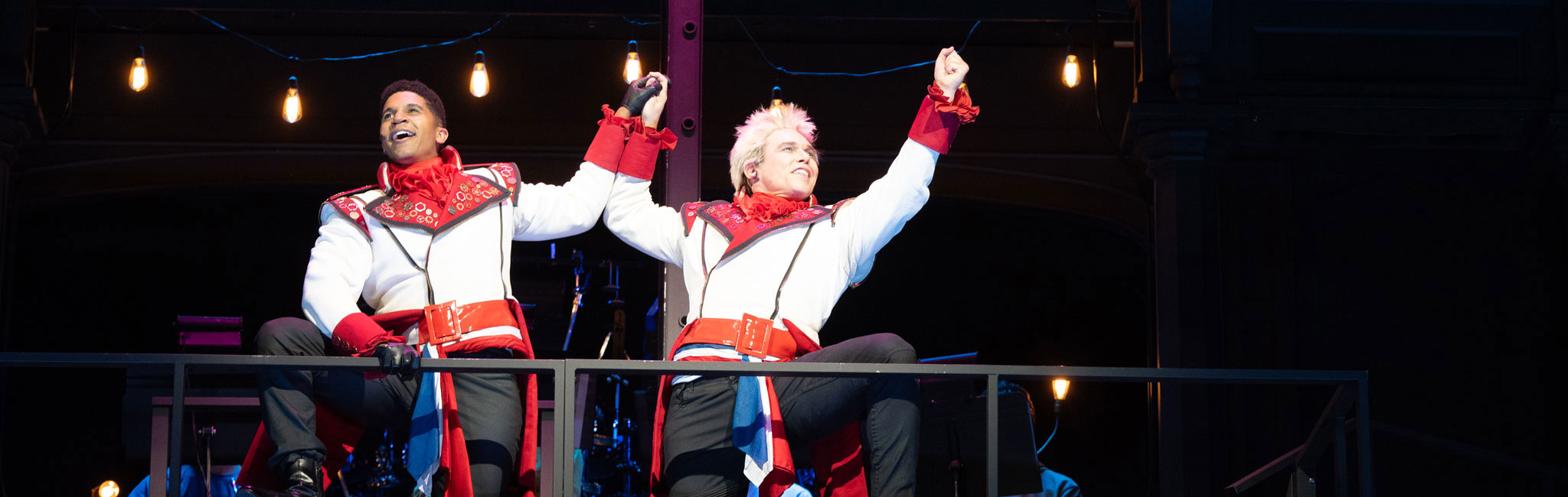 Two performers in matching costumes raising fists in triumph on stage balcony.